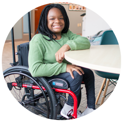 Young black girl with braids wearing a sweater and jean using a manual wheelchair smiling at the camera.
